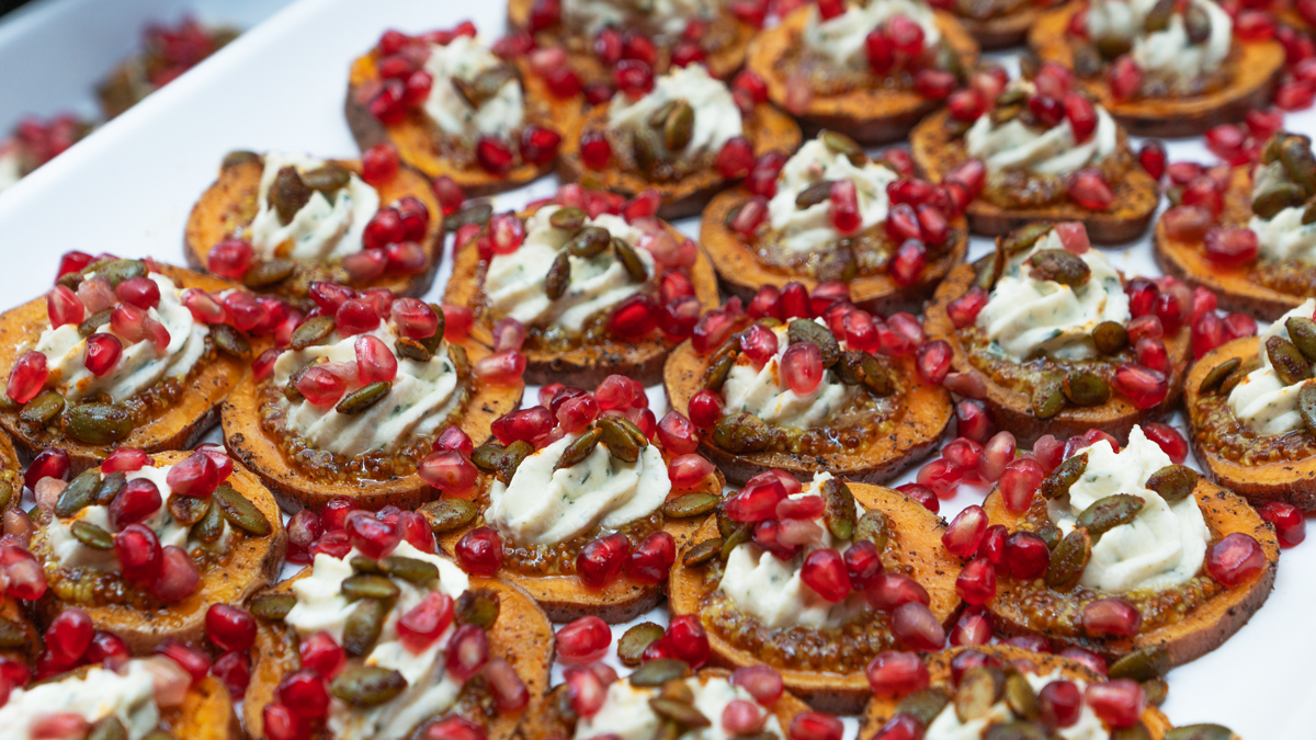 Image is a close up of sweet potato slices topped with a cream sauce, pumpkin seeds, and pomegranate seeds