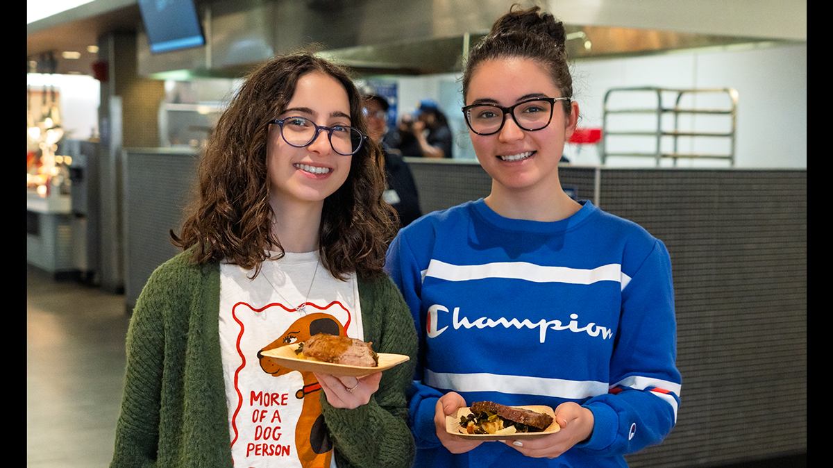 Image shows two young women with brown hair and glasses holding plates of food.