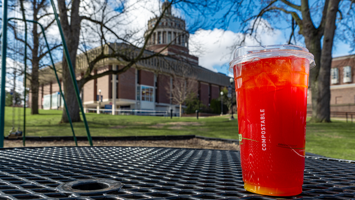 The image shows a red cold drink with ice on a table in front of the Rush Rhees Library.