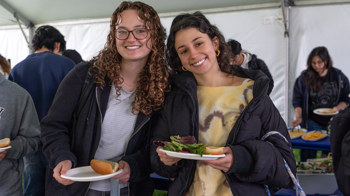 Image shows two young women smiling and looking at the camera. One is holding a plate with an empty hotdog bun and the other is holding a plate with salad and a hamburger bun.