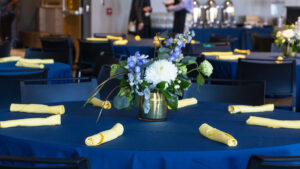 Image shows a table with flowers in the center, a blue tablecloth, and yellow napkins.
