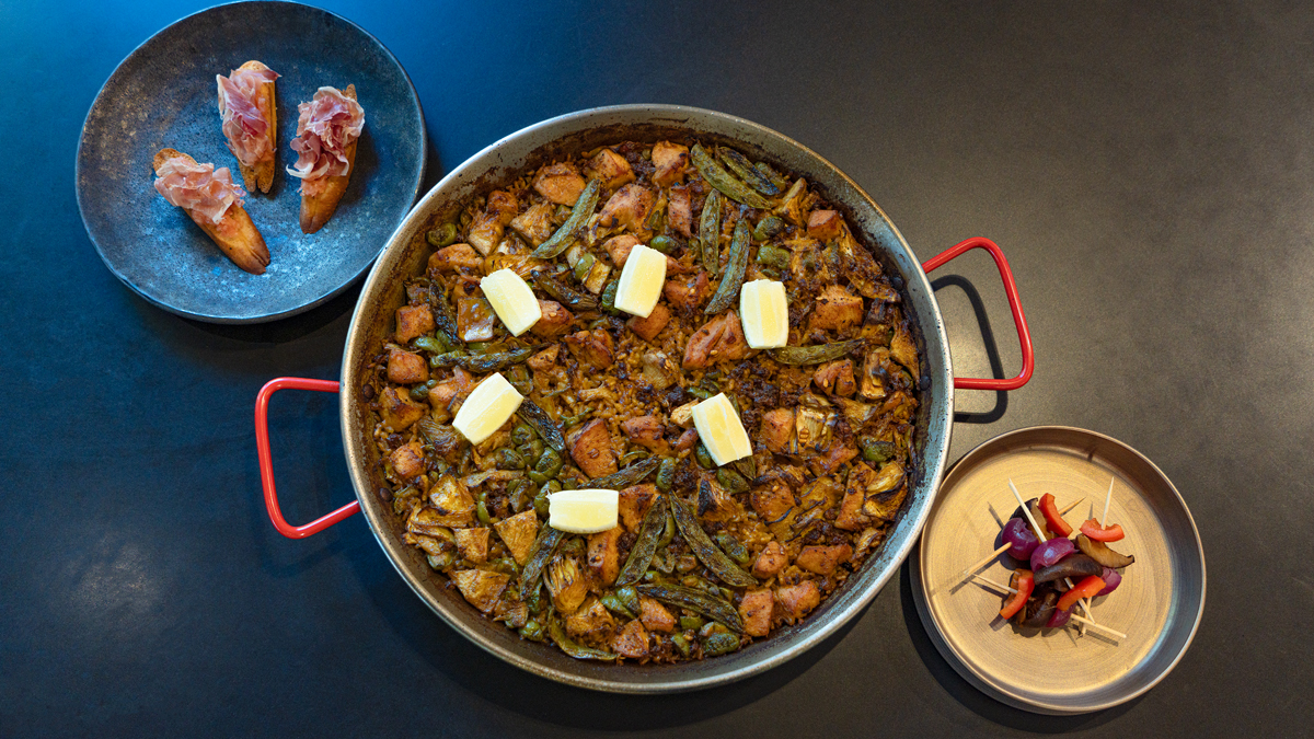 Image shows a large pan of paella with two small side dishes, one with toast topped with prociutto and the other with skewered vegetables.