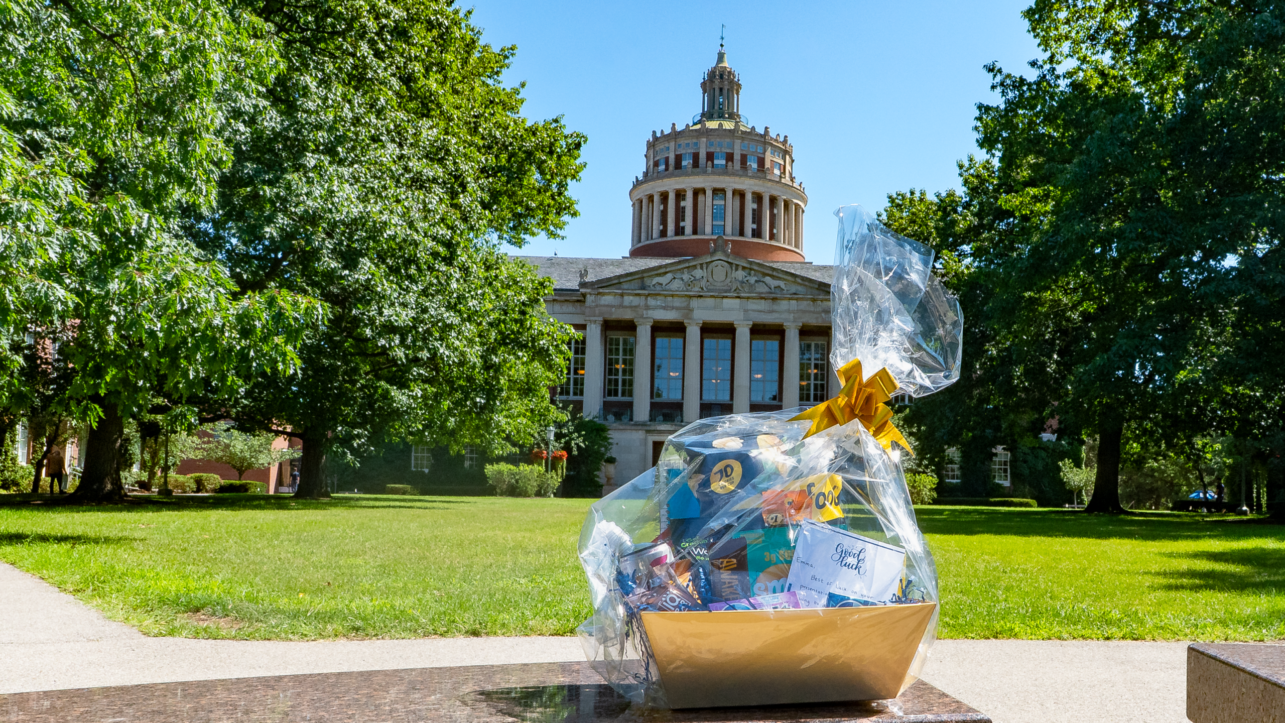 The image displays a care package resting on a bench, with Rush Rees library towering over in the background.